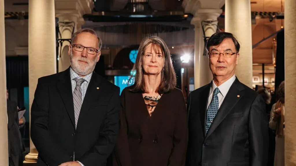 Three formally-dressed people stand in a classical hall, flanked by pillars behind them.